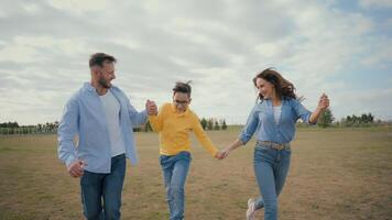 Healthy Caucasian happy family joyful woman female mom mother holding hands dad male man little boy child son kid jumping run playing running jump together outdoors nature park. Active rest childhood video