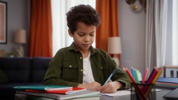 Concentrated little African American boy son kid child pupil schoolboy at home table writing task homework write study distant education learning positive studying learn smiling looking at camera video