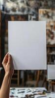 a woman hand holding up a a blank white piece of A4 paper, there is an art studio in the background, realistic photo