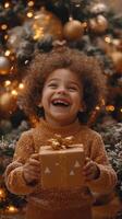 A joyful child with curly hair smiles brightly while holding a gift in front of a Christmas tree, adorned with warm lights and decorations. photo