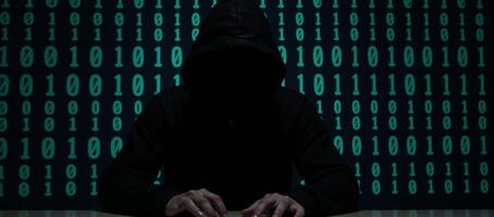 A hooded criminal silhouetted while hacking on binary code background cybercrime concept photo