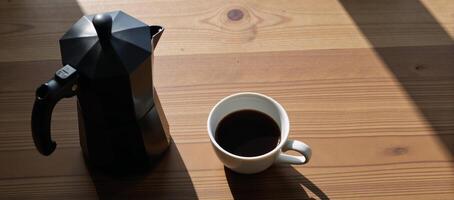 A black coffee pot stands beside a white coffee cup on a wooden table photo