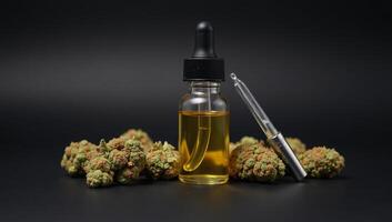 A small glass bottle containing cannabis oil alongside a pipette surrounded by dried marijuana flowers against a black backdrop photo