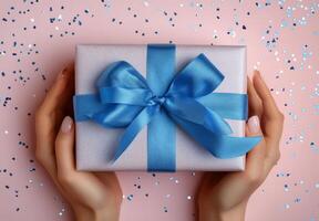 Hands holding a beautifully wrapped blue gift box with a satin ribbon on a light blue background photo