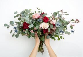 Two hands hold a pink and red rose arrangement with greenery against a neutral background photo