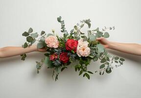 Two hands hold a pink and red rose arrangement with greenery against a neutral background photo