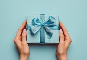 Hands holding a beautifully wrapped blue gift box with a satin ribbon on a light blue background photo