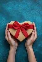 A person holds a heart shaped gift box tied with a red ribbon against a teal background photo