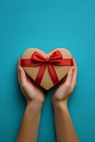 A person holds a heart shaped gift box tied with a red ribbon against a teal background photo