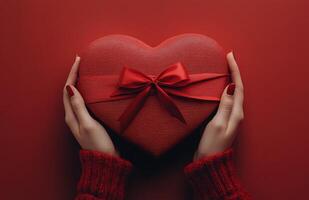 Hands presenting a heart shaped gift box tied with a ribbon against a red background photo