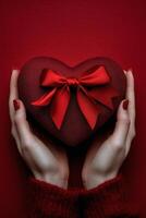 A person holds a heart shaped gift box tied with a red ribbon against a teal background photo