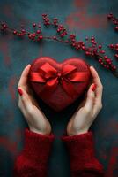 A person holds a heart shaped gift box tied with a red ribbon against a teal background photo