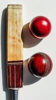 cricket bat lies next to two red cricket balls on a white background highlighting the wood grain and leather texture under soft shadows in midday lighting. photo