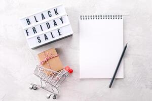 Flat lay of lightbox with text black friday sale, grocery cart with gift present, notebook with place for text photo