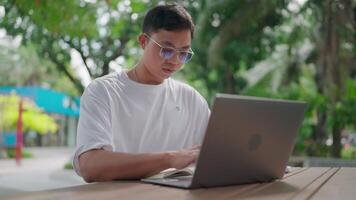 Young Asian man with glasses using laptop while sitting at table outdoors working intently in public park. Natural environment. Learning. Typing on keyboard. Technology. Internet. Taking notes. video