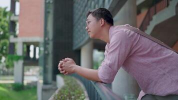Student standing with bag and smiling. Man wearing glasses and plaid shirt standing with bag, leaning against railing of balcony on walkway around university building, looking relaxed and happy. video