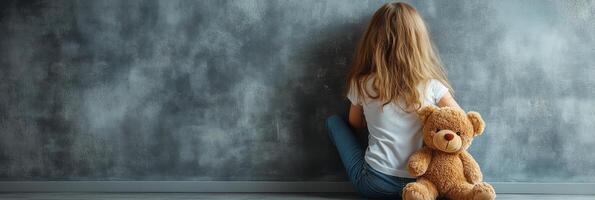 A young child sits alone on the floor with a teddy bear in a softly lit room photo