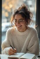 Young woman in cozy sweater writing in notebook by sunny window in a coffee shop photo