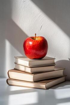 A vibrant red apple resting atop three stacked books on a sunny surface with shadows photo