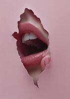 A close up view of a mouth breaking through pink paper with glossy lips and white teeth photo