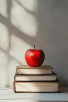 A vibrant red apple resting atop three stacked books on a sunny surface with shadows photo
