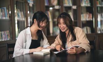 Two women are sitting at a table in a library, looking at a book photo