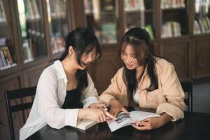 Two women are sitting at a table in a library, looking at a book photo