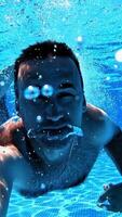 Man swims with underwater camera inside the swimming pool. Transparent water background of a pool and male adult swimming underwater and looking into camera. Vertical video
