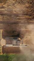 Combine harvester working in the field and producing clouds of dust rising in the air. Drone footage over the machine gathering crops. Vertical video