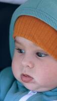 Lovely Caucasian boy with sweet cheeks sits in stroller. Cute child wearing warm clothes and cap having a walk in pram. Close up. Vertical video