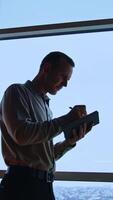 Busy man taking notes into paper notebook. Male worker stands in front of big window and writes in his book. Low angle view. Vertical video