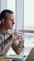 Calm relaxed man sit in front of his computer with a cup in hands. Thoughtful man looks into window and then straight ahead. Vertical video