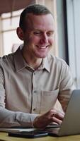 Smiling young man working at his laptop. Male employee says something and looks into window. Blurred backdrop. Vertical video