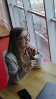 Long-haired young woman having tea sitting at the table. Lady enjoying lunch break and looking out of the window. High angle view. Vertical video