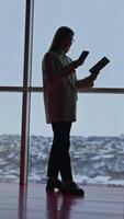 Lady in suit speaks on the phone and looks at the notebook. Full-length portrait of a woman standing in front of panoramic windows at cityscape backdrop. Vertical video