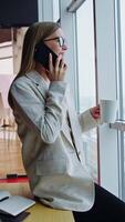 Resilient lady sits on the desk, speaks on the phone and holds a cup. Lunch break in office with big windows. Cityscape at backdrop. Vertical video