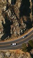 Cars running quickly by the two-way road in the mountains. High rocks covered with moss and rare bushes on sunny day from top view. Vertical video