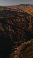 Mountainous territory with little greenery at sunset. Motorway passing through the rocks with numerous cars. Aerial view. Vertical video