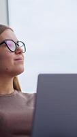 Confident relaxed blonde woman working on computer. Female looks into window for a moment and continues her work smiling. Low angle view. Vertical video