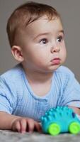Beautiful baby lying on belly on the floor with toy in front of him. Cute child looks up and smiles to somebody adorably. Blurred backdrop. Vertical video