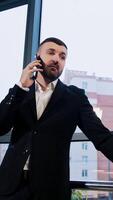 Man having unpleasant phone talk. Businessman in suit stands at panoramic window speaking on the phone. Low angle view. Vertical video