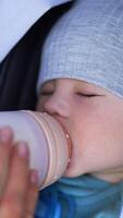Mother is feeding her little son from a bottle. Lovely child eats lying in a stroller and falling asleep. Close up. Vertical video