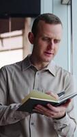 Young man leaned on the window holding a telephone and a paper notebook. Man turning pages in a book and comments something. Vertical video
