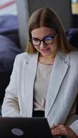 Lady in light jacket sitting in bean bag chair working on her laptop. Comfortable workplace in a nice modern creative office. Blurred backdrop. Vertical video