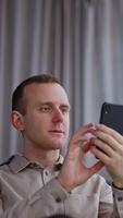 Smiling young man looks at the phone in front of him. Male employee relaxing at lunch time. Grey backdrop in blur. Vertical video