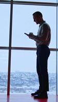 Office employee at the window with phone and white cup. Full-length portrait of a slim man at the backdrop of panoramic window. Vertical video