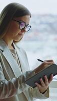 Young woman taking notes in her book standing at the window. Lady in light jacket looks thoughtfully into the blurred window. Vertical video