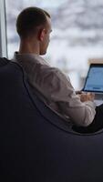 Busy male freelancer sits in bean bag chair with his back to camera. Man works on a laptop with graphs at the screen. Window with blurred cityscape at backdrop. Vertical video