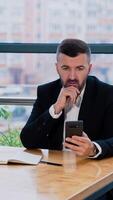 Thoughtful businessman focused on the screen of his smartphone. Busy man scrolling information in his phone sitting at the desk. Blurred backdrop. Vertical video