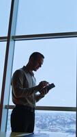Handsome slim man stands at the window looking into his notebook. Office worker takes some notes. Cityscape at backdrop. Vertical video